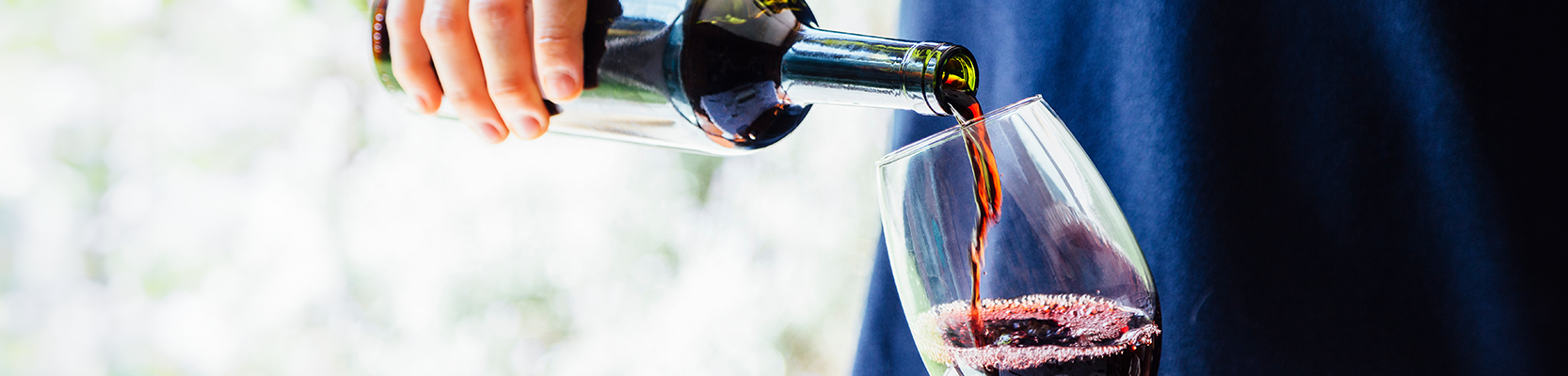 Rotwein der aus der Flasche in ein Glas gegossen wird. Die Person trägt ein blaues Shirt